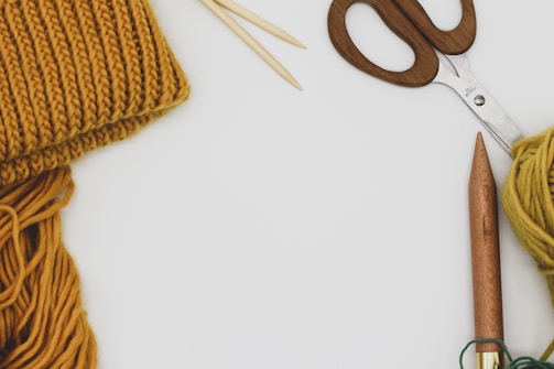 Knitting materials and tools are arranged on a white surface. A pair of wooden-handled scissors, a ball of mustard-colored yarn, knitting needles, and a partially knitted piece are visible.