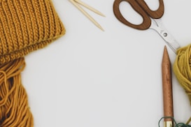Knitting materials and tools are arranged on a white surface. A pair of wooden-handled scissors, a ball of mustard-colored yarn, knitting needles, and a partially knitted piece are visible.
