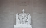 A large marble statue of a seated man surrounded by a grand architectural setting. The figure is dressed in historical attire and is portrayed with a solemn expression. The backdrop features an engraved text commemorating his contributions.