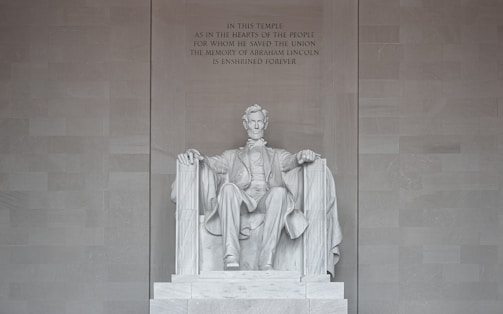 A large marble statue of a seated man surrounded by a grand architectural setting. The figure is dressed in historical attire and is portrayed with a solemn expression. The backdrop features an engraved text commemorating his contributions.