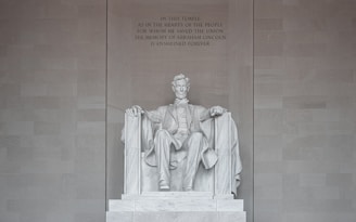 A large marble statue of a seated man surrounded by a grand architectural setting. The figure is dressed in historical attire and is portrayed with a solemn expression. The backdrop features an engraved text commemorating his contributions.
