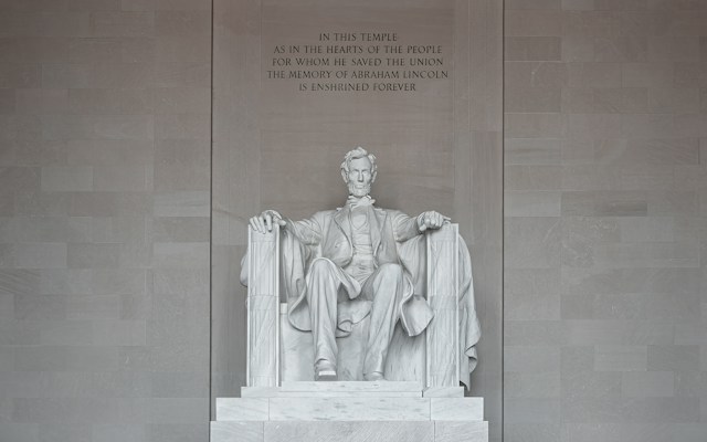 A large marble statue of a seated man surrounded by a grand architectural setting. The figure is dressed in historical attire and is portrayed with a solemn expression. The backdrop features an engraved text commemorating his contributions.