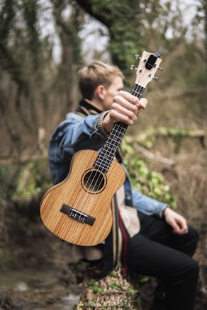 A person sitting outdoors in a natural setting, holding up a wooden ukulele. The surroundings are lush with greenery and the atmosphere appears serene. The focus is on the ukulele being prominently displayed by the individual on their lap, dressed in casual attire with a denim jacket.