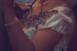 An elegant off-shoulder garment adorned with intricate blue jewel-like embellishments. The fabric has a shiny, silky appearance, giving it a luxurious feel. The accessories include a matching necklace and bracelet, contributing to the overall ornate and sophisticated look.