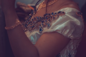 An elegant off-shoulder garment adorned with intricate blue jewel-like embellishments. The fabric has a shiny, silky appearance, giving it a luxurious feel. The accessories include a matching necklace and bracelet, contributing to the overall ornate and sophisticated look.