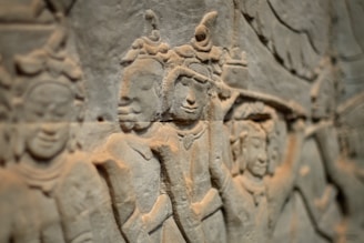 Stone relief carving featuring multiple figures in traditional attire, possibly depicting a historical or religious scene. The style suggests ancient artistry with intricate detailing in the headdresses and robes.