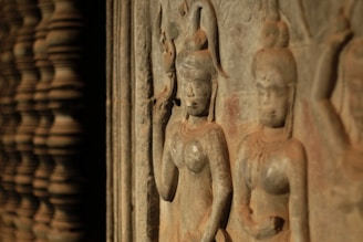 Ancient stone carvings depict two figures with intricate hairstyles and traditional attire, showcasing detailed craftsmanship. The carvings appear aged, with a warm patina hinting at the passage of time.