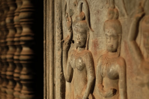 Ancient stone carvings depict two figures with intricate hairstyles and traditional attire, showcasing detailed craftsmanship. The carvings appear aged, with a warm patina hinting at the passage of time.