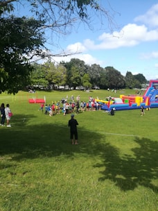 A group of people, including children and adults, are gathered on a grassy field next to a colorful inflatable obstacle course. The area is surrounded by trees under a partly cloudy sky, while additional play equipment and people are visible in the background.