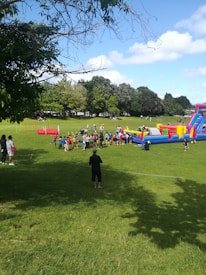 A group of people, including children and adults, are gathered on a grassy field next to a colorful inflatable obstacle course. The area is surrounded by trees under a partly cloudy sky, while additional play equipment and people are visible in the background.