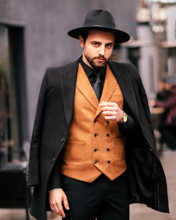 A man with a beard wearing a stylish, wide-brimmed hat and a black coat draped over his shoulders stands confidently. He is dressed in a brown, double-breasted vest paired with a black shirt and tie. The background is blurred, giving a sense of an urban setting.