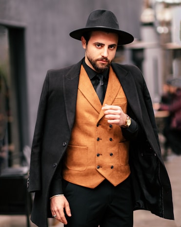 A man with a beard wearing a stylish, wide-brimmed hat and a black coat draped over his shoulders stands confidently. He is dressed in a brown, double-breasted vest paired with a black shirt and tie. The background is blurred, giving a sense of an urban setting.