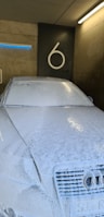 A car is completely covered in a layer of white soap foam inside a wash bay. The background features a concrete wall with the number 6 prominently displayed. Overhead lighting casts reflections on the soapy surface of the vehicle.