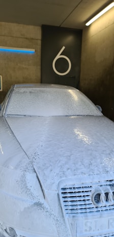 A car is completely covered in a layer of white soap foam inside a wash bay. The background features a concrete wall with the number 6 prominently displayed. Overhead lighting casts reflections on the soapy surface of the vehicle.