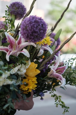 A vibrant bouquet of flowers featuring a variety of blooms including large purple alliums, pink and white lilies, bright yellow roses, and small yellow daisies. The bouquet is lush with greenery and other small white flowers, creating a rich and colorful arrangement.