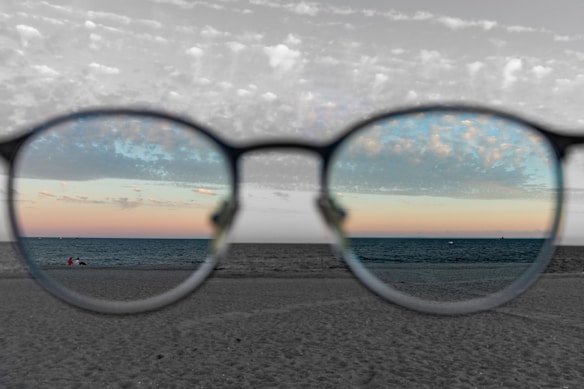 An artistic photograph showing a beach scene viewed through eyeglasses. The area within the lenses is in color, displaying a serene sunset over the ocean, while the rest of the photograph is in black and white. A person is seen sitting or lying on the beach.