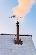 A snowy rooftop with a chimney is seen with smoke rising into a clear blue sky. Mounted on top of the chimney is a decorative weather vane featuring a silhouette of a cat.
