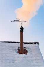 A snowy rooftop with a chimney is seen with smoke rising into a clear blue sky. Mounted on top of the chimney is a decorative weather vane featuring a silhouette of a cat.