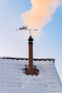 A snowy rooftop with a chimney is seen with smoke rising into a clear blue sky. Mounted on top of the chimney is a decorative weather vane featuring a silhouette of a cat.