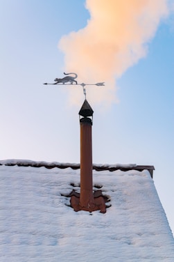A snowy rooftop with a chimney is seen with smoke rising into a clear blue sky. Mounted on top of the chimney is a decorative weather vane featuring a silhouette of a cat.