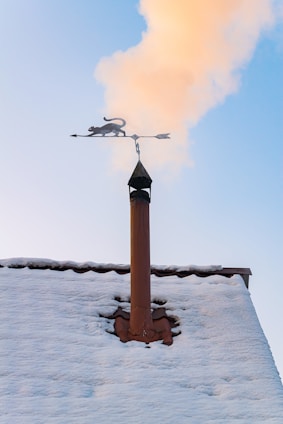 A snowy rooftop with a chimney is seen with smoke rising into a clear blue sky. Mounted on top of the chimney is a decorative weather vane featuring a silhouette of a cat.