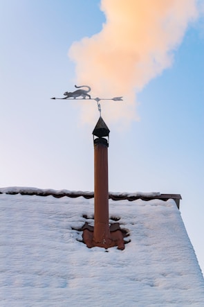 A snowy rooftop with a chimney is seen with smoke rising into a clear blue sky. Mounted on top of the chimney is a decorative weather vane featuring a silhouette of a cat.
