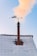 A snowy rooftop with a chimney is seen with smoke rising into a clear blue sky. Mounted on top of the chimney is a decorative weather vane featuring a silhouette of a cat.