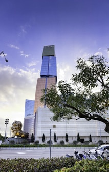 A tall, modern skyscraper with the logo at the top stands prominently against a blue sky with scattered clouds. The building features a combination of reflective glass and golden panels. In the foreground, a large golden lion sculpture is positioned to the left. A lush tree partially frames the right side of the scene, with a few parked motorcycles visible below.
