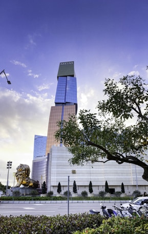 A tall, modern skyscraper with the logo at the top stands prominently against a blue sky with scattered clouds. The building features a combination of reflective glass and golden panels. In the foreground, a large golden lion sculpture is positioned to the left. A lush tree partially frames the right side of the scene, with a few parked motorcycles visible below.