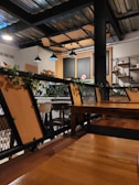 A modern cafe interior with industrial design elements. Wooden tables and metal chairs are arranged neatly, surrounded by green plants on railings. Multiple hanging lights illuminate the space, casting a warm glow. The walls have shelving units with decorative items, and a no smoking sign is visible.
