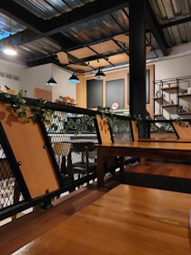 A modern cafe interior with industrial design elements. Wooden tables and metal chairs are arranged neatly, surrounded by green plants on railings. Multiple hanging lights illuminate the space, casting a warm glow. The walls have shelving units with decorative items, and a no smoking sign is visible.