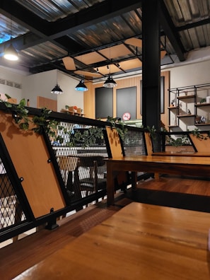 A modern cafe interior with industrial design elements. Wooden tables and metal chairs are arranged neatly, surrounded by green plants on railings. Multiple hanging lights illuminate the space, casting a warm glow. The walls have shelving units with decorative items, and a no smoking sign is visible.