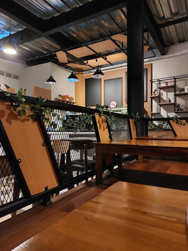 A modern cafe interior with industrial design elements. Wooden tables and metal chairs are arranged neatly, surrounded by green plants on railings. Multiple hanging lights illuminate the space, casting a warm glow. The walls have shelving units with decorative items, and a no smoking sign is visible.