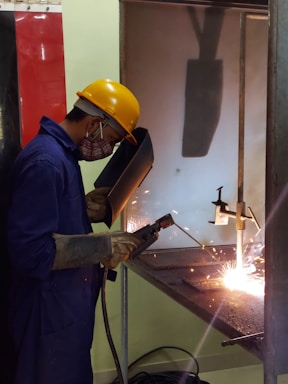 A worker engaged in welding, wearing a yellow safety helmet, protective gloves, and a face shield. Sparks are visible as the welding process takes place inside a workshop. The worker is standing at a metal workbench, carefully operating a welding tool.