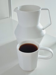A minimalistic setup featuring a white ceramic pitcher and a matching white mug filled with dark coffee on a white surface.