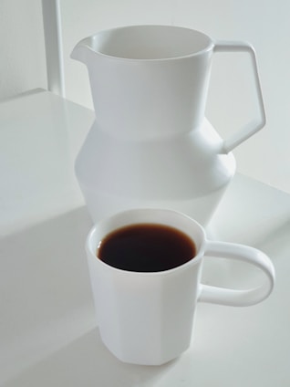 A minimalistic setup featuring a white ceramic pitcher and a matching white mug filled with dark coffee on a white surface.