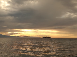 A cargo ship sailing across calm blue waters with the sun setting in the background.