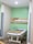 A hospital room with a bed on wheels. The bed is neatly made with white sheets and a brown blanket. The room has pale green walls, a wooden panel with medical equipment including electrical outlets and an IV stand. There are curtains partially drawn on both sides of the bed for privacy, and the flooring is tiled.