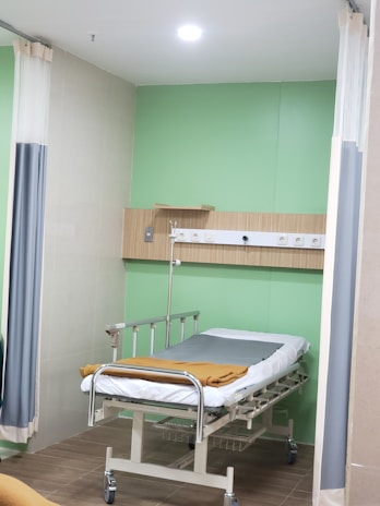 A hospital room with a bed on wheels. The bed is neatly made with white sheets and a brown blanket. The room has pale green walls, a wooden panel with medical equipment including electrical outlets and an IV stand. There are curtains partially drawn on both sides of the bed for privacy, and the flooring is tiled.