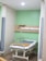 A hospital room with a bed on wheels. The bed is neatly made with white sheets and a brown blanket. The room has pale green walls, a wooden panel with medical equipment including electrical outlets and an IV stand. There are curtains partially drawn on both sides of the bed for privacy, and the flooring is tiled.