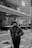 A person wearing a puffer jacket and beanie stands in an urban setting with modern buildings in the background. The scene is monochromatic, suggesting it might be cold, with some snow visible on the ground. Other people are present in the distance, contributing to a city atmosphere.