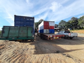 A scene featuring several trucks parked on a dirt ground under a clear blue sky. The trucks have large containers, with one painted in blue and another in red, white, and blue. A pickup truck can be seen beside the larger vehicles. Trees and a small building are visible in the background.