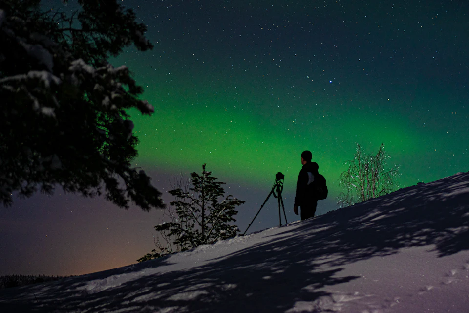 A photographer waits beside a camera on a tripod under a green aurora.