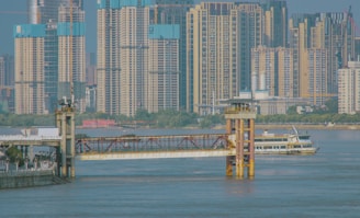 A busy urban riverfront scene with high-rise buildings in the background. The river is populated with a large ferry boat near a dock and modern infrastructure including bridges and piers.
