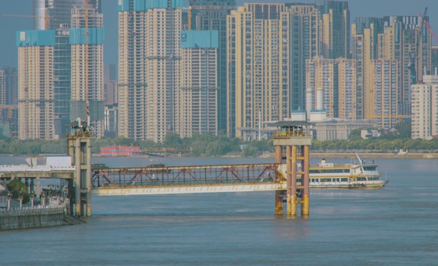 A busy urban riverfront scene with high-rise buildings in the background. The river is populated with a large ferry boat near a dock and modern infrastructure including bridges and piers.
