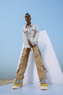 A person wearing a checkered jacket and beige cargo pants stands confidently on a white platform. The shoes feature a colorful design incorporating blue, yellow, and white. Sunglasses adorn the face, and the background is a clear blue sky, creating a minimalistic and modern aesthetic.