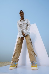 A person wearing a checkered jacket and beige cargo pants stands confidently on a white platform. The shoes feature a colorful design incorporating blue, yellow, and white. Sunglasses adorn the face, and the background is a clear blue sky, creating a minimalistic and modern aesthetic.
