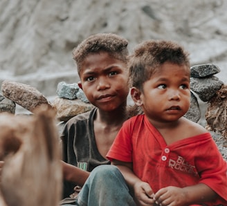 Two children sit closely together, surrounded by a stack of stones. The older child wears a dark shirt, while the younger one is in a bright red shirt. Their expressions convey curiosity and seriousness.