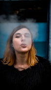 A person with light brown hair is exhaling smoke in front of a blue background while wearing a black garment.