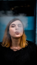 A person with light brown hair is exhaling smoke in front of a blue background while wearing a black garment.